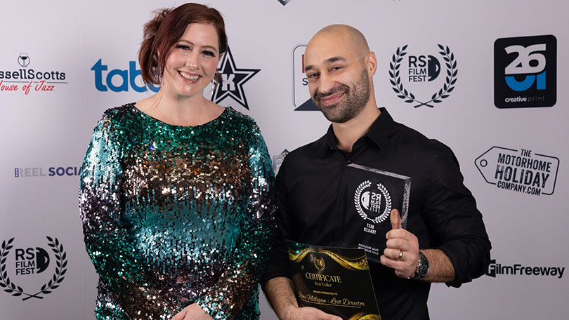 Samuel Lodato and Sophie Slavin pose together at the RS Film Fest. Samuel holds a trophy and certificate for Best Trailer, while Sophie, the festival director, smiles beside him. They stand in front of a backdrop with various sponsor logos and laurels.