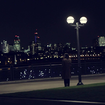 Film Still. A long-haired man stands in his trench coat by a streetlight, taking in the London cityscape.