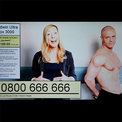 Film still. A young blond woman on a teleshopping channel. A muscular man stands next to her, posing with his top off.
