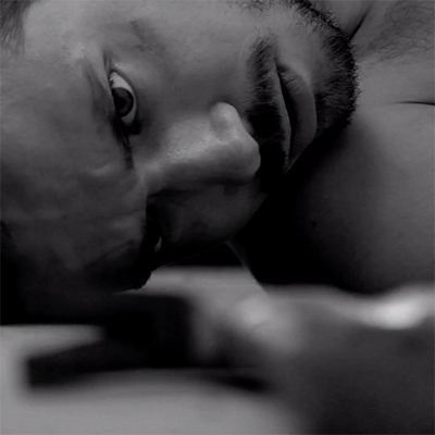 Film Still. Moody black & white. Close-up of a man, lying on his side, eyes fixed on something on the ground next to his face.