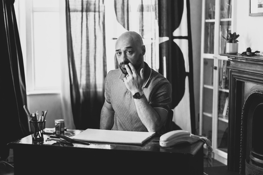 A glossy black and white picture of a bearded bald man sitting at a desk of a nicely furnished room.