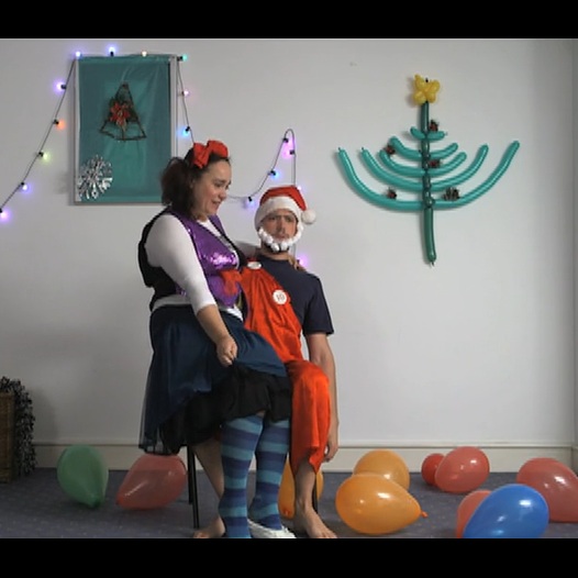 An eccentrically-dressed grown woman sits on the lap of a badly-dressed Santa Clause. There are colourful balloons by their feet but their surroundings are otherwise stark.